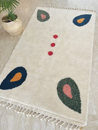 Full shot of a Beni Ourain Moroccan Rug created in collaboration with White Otter Design Co. It features a cream base with green and blue petals, and red dots. On a marble floor with a plant in frame.