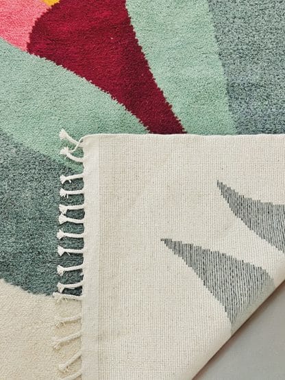 Close up of the folded corner of a hand-knotted Beni Ourain style rug created with White Otter Design Co. It features a cream base with a large floral design in green, red, pink and yellow.