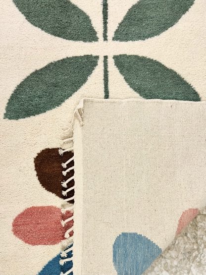 Close up of the folded corner of a hand-knotted Beni Ourain style rug created with White Otter Design Co. It features a cream base with a large floral design in green, red, pink and yellow.
